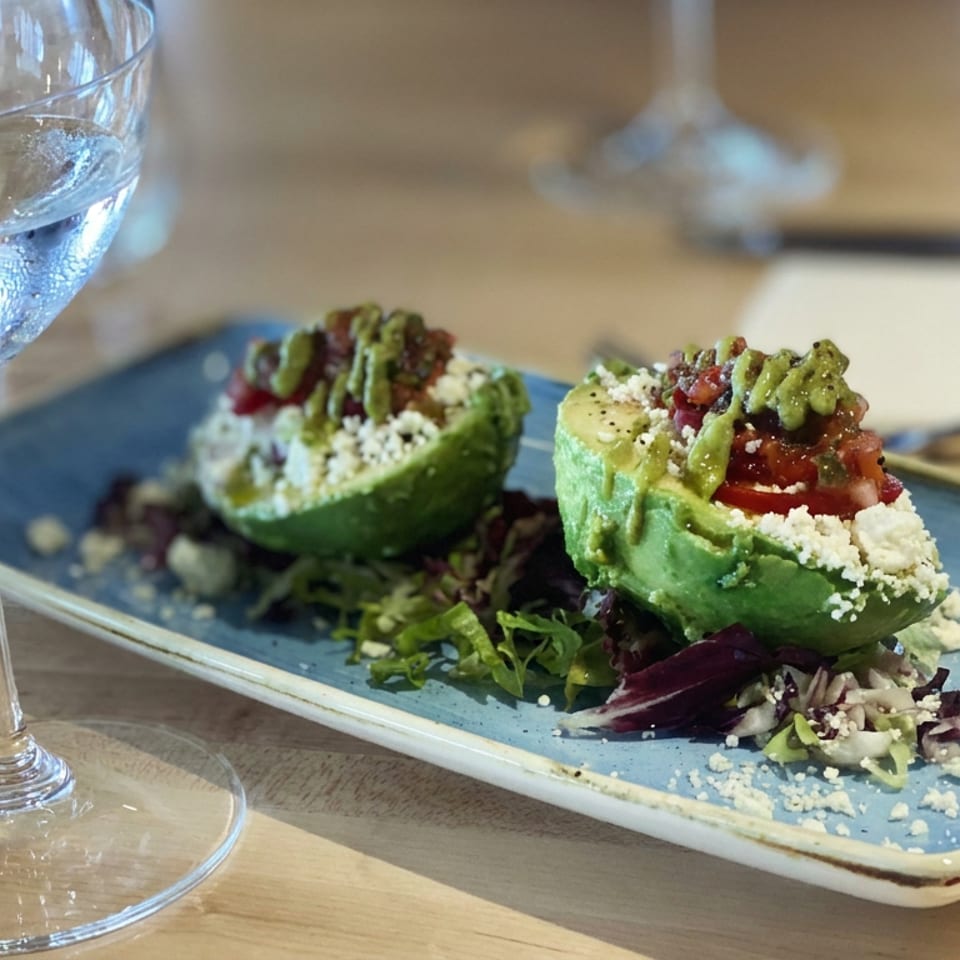 Two avocado halves filled with tomato, greens, and crumbled cheese on a blue plate beside a glass of white wine.