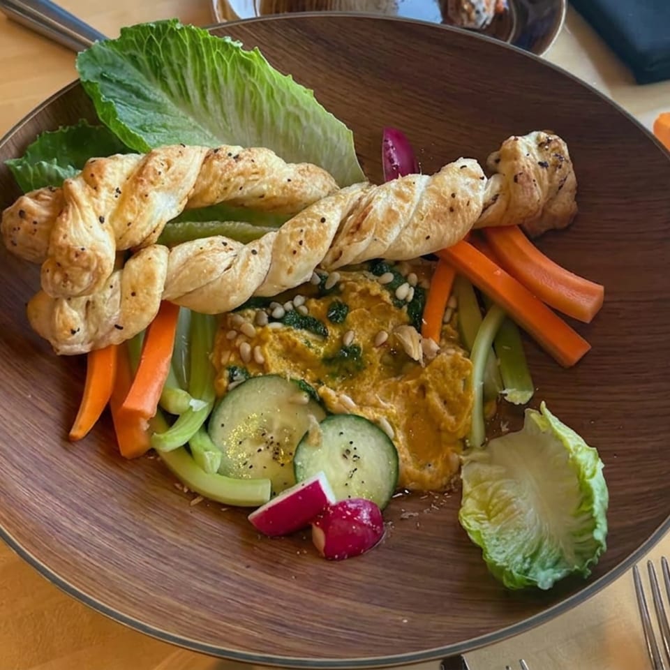 Plated hummus with twisted flatbread, lettuce, cucumber, carrots, celery, radishes, and pine nuts on a wooden table.