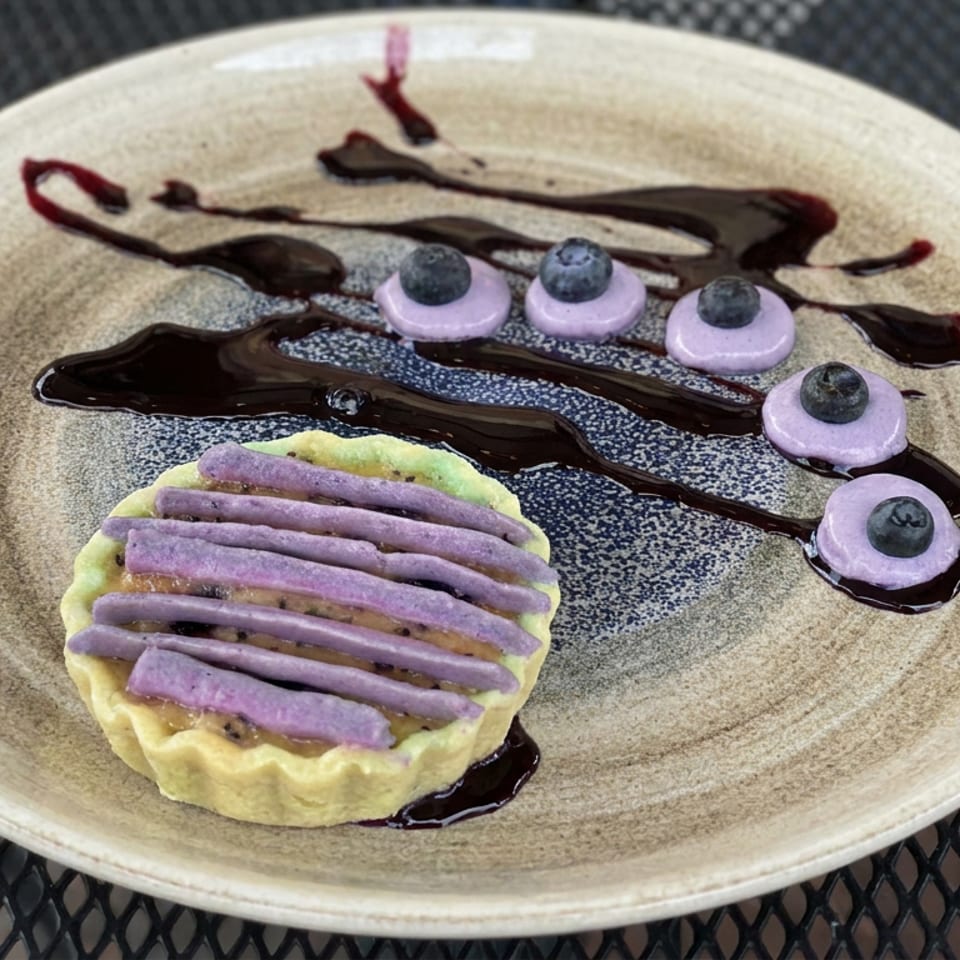 Dessert tart topped with purple cream, blueberries, and chocolate sauce on a ceramic plate atop a metal table.