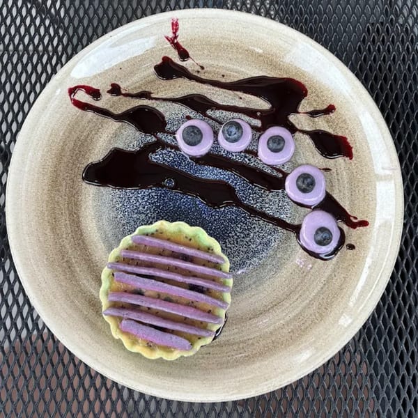 A dessert plate with a small tart and five blueberry-topped purple circles, drizzled with dark sauce.
