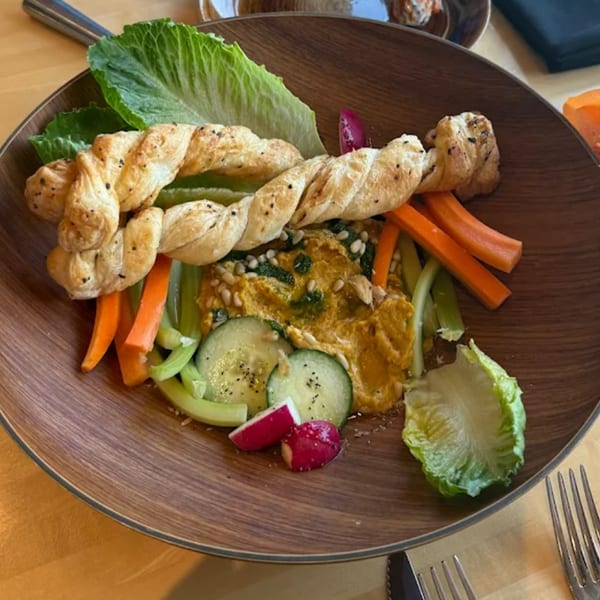 A wooden bowl of hummus with pine nuts, pita bread, carrots, celery, cucumbers, and radishes.