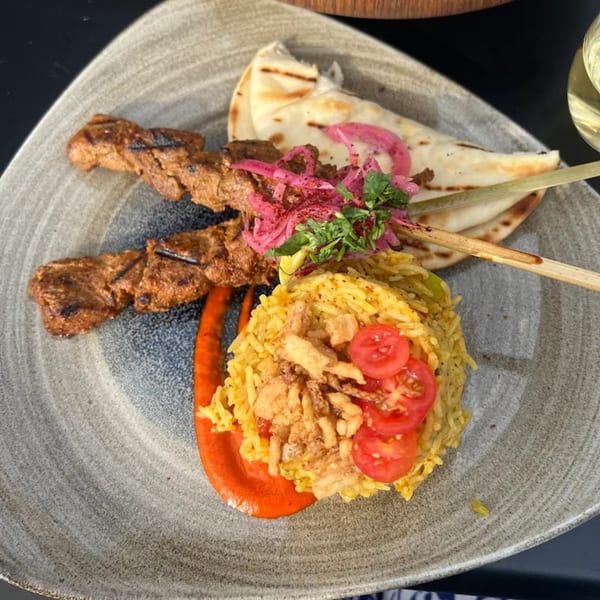 Grilled meat skewers, yellow rice with tomatoes, red sauce, and flatbread with pickled onions on a plate.
