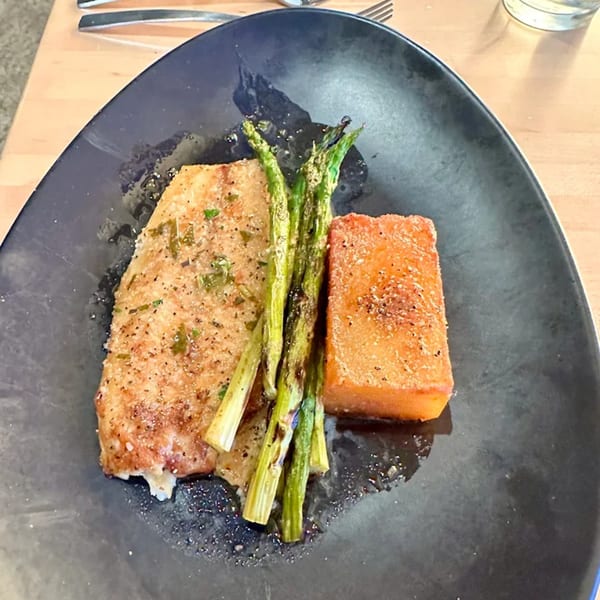 Mediterranean dish with a golden fish fillet, grilled asparagus, and a sweet potato block on a dark plate.