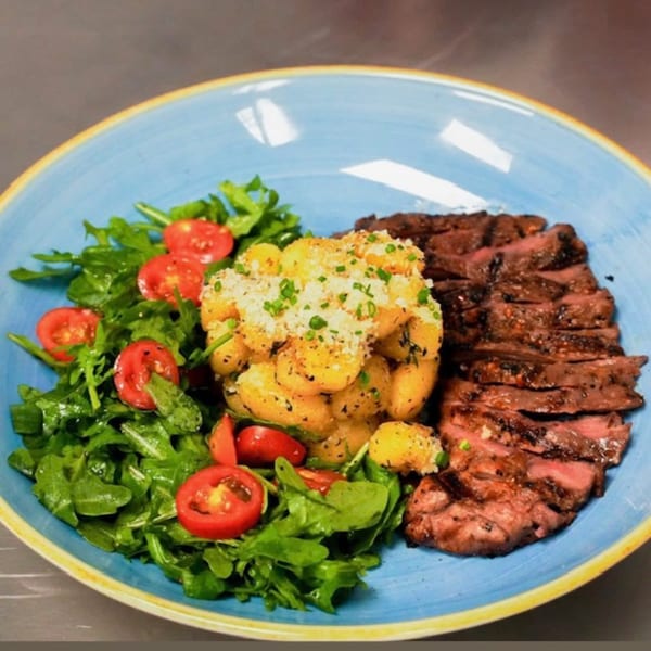 Grilled steak slices, gnocchi, and a fresh arugula salad with cherry tomatoes on a blue plate.