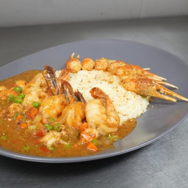 Shrimp and grits with a rich sauce, vegetables, and shrimp skewers on a gray plate.