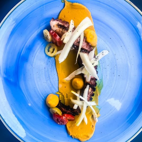 Grilled octopus with yellow puree, white vegetables, and cherry tomatoes on a blue plate.