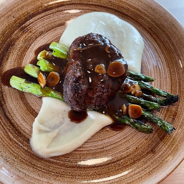 A Mediterranean dish with steak, asparagus, mashed potatoes, and mushroom gravy on a brown plate.
