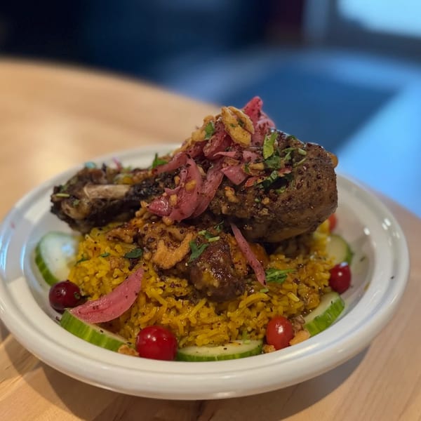 A Mediterranean dish with a large piece of meat, yellow rice, pickled onions, and cucumber slices.